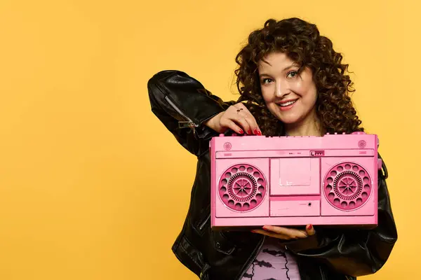 Woman with curly hair smiles, holding pink boombox against yellow background, adorned in black leather jacket. — Stock Photo