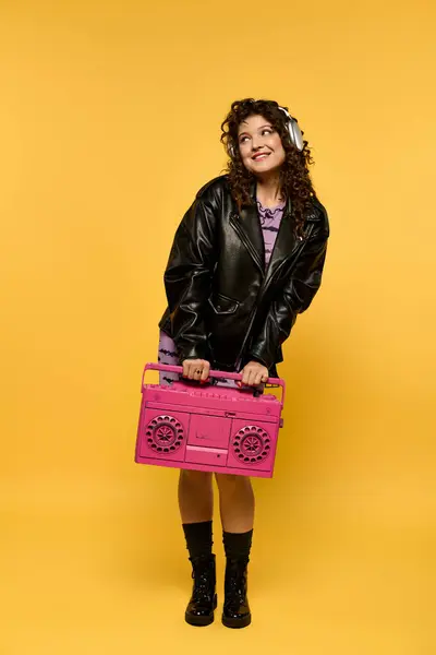 A young woman with curly hair poses in a black leather jacket while holding a pink boombox against a yellow background. — Stock Photo