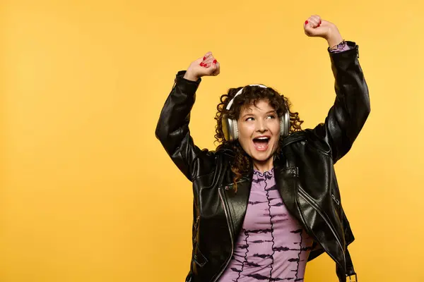 Eine stylische junge Frau mit lockigem Haar, schwarzer Lederjacke und Kopfhörern tanzt aufgeregt vor gelbem Hintergrund. — Stockfoto