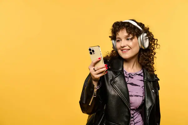 A young woman with curly hair and a leather jacket smiles while looking at her smartphone against a bright yellow background. — Stock Photo