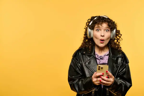 A young woman in a leather jacket with curly hair poses in front of a yellow background with a surprised expression. — Stock Photo