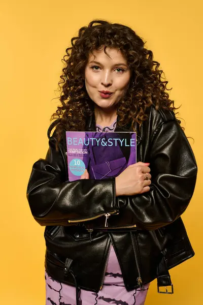 A young woman with curly hair wears a black leather jacket while posing against a yellow background. She is holding a fashion magazine. — Stock Photo