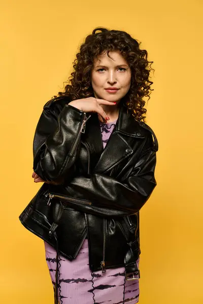 A young woman with curly hair is wearing a black leather jacket against a vibrant yellow background. — Stock Photo