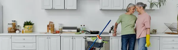A mature lesbian couple cleans their modern kitchen together, smiling and leaning in towards each other. — Stock Photo