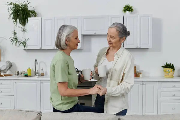 Due donne mature, probabilmente una coppia lesbica, condividono un momento di tranquillità nella loro cucina moderna, abbracciando tazze di caffè e sorridendosi. — Foto stock