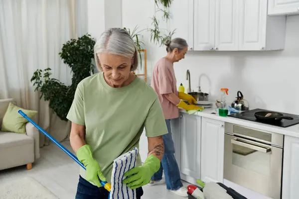 Two mature lesbians tidy their apartment. One mops while the other washes dishes. — Stock Photo
