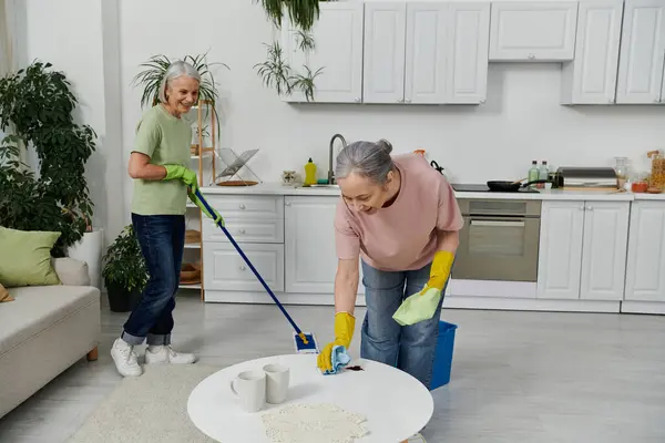 A lesbian couple cleans their modern apartment together. One woman mops the floor, while the other wipes down a table. — Stock Photo