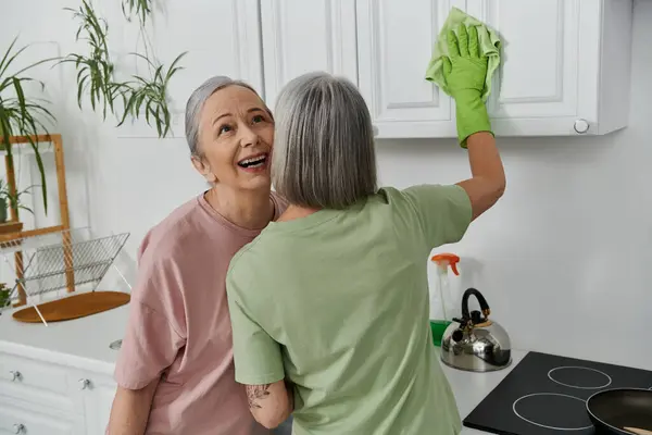 Un couple de lesbiennes nettoie leur cuisine moderne ensemble, une femme essuyant les armoires tandis que l'autre regarde et sourit. — Photo de stock