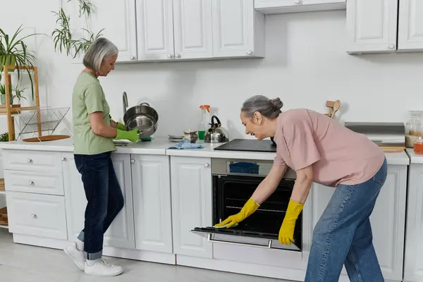 Ein reifes lesbisches Paar putzt gemeinsam seine moderne Wohnung. Eine Frau spült Geschirr, während die andere den Ofen putzt. — Stockfoto