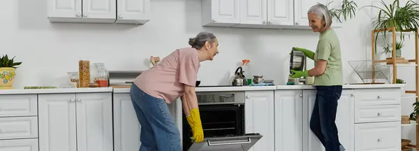 Un couple de lesbiennes mûres nettoie leur cuisine moderne ensemble, une femme nettoyant le four tandis que l'autre lave la vaisselle. — Photo de stock