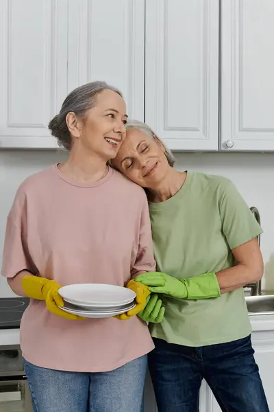 An older lesbian couple cleans their modern apartment together. The woman in the pink shirt holds a stack of dishes. — Stock Photo