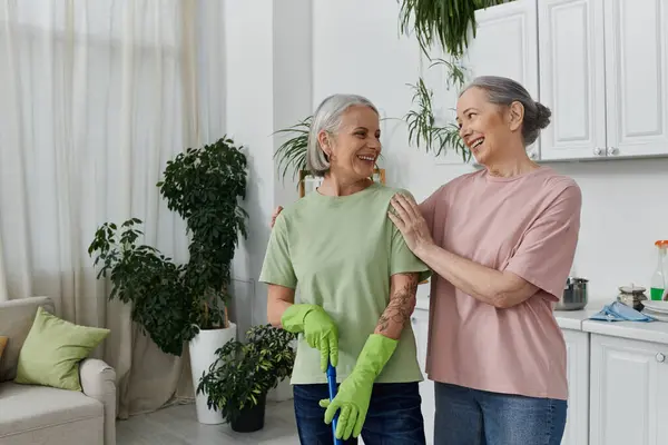 Duas mulheres maduras sorriem uma para a outra enquanto limpam seu apartamento moderno. — Fotografia de Stock