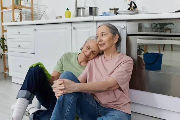 Um casal lésbico mais velho faz uma pausa da limpeza de sua cozinha moderna, encostado aos armários. — Fotografia de Stock