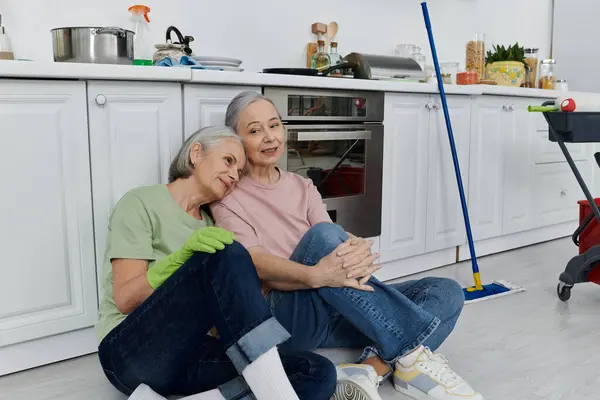 A mature lesbian couple takes a break from cleaning their modern kitchen, with one woman resting her head on the others shoulder. — Stock Photo
