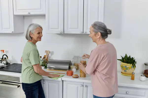 Two mature women, likely a lesbian couple, clean their modern kitchen together. One woman wipes the counter while the other sorts items. — Stock Photo