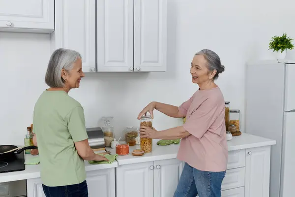 Um casal de lésbicas limpa sua cozinha moderna apartamento, uma mulher organizando itens de despensa, enquanto o outro limpa o balcão. — Fotografia de Stock