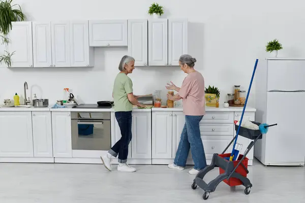 A mature lesbian couple cleans their modern kitchen, one woman wiping the counters while the other organizes supplies. — Stock Photo