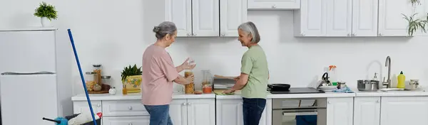 Um casal de lésbicas mais velhas limpa a sua cozinha moderna juntos. Uma mulher segura um frasco enquanto a outra prepara uma refeição na bancada.. — Fotografia de Stock
