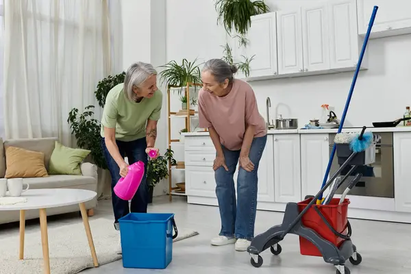 Dois parceiros lésbicas limpam seu apartamento moderno juntos. Uma mulher derrama solução de limpeza em um balde, enquanto os outros relógios. — Fotografia de Stock