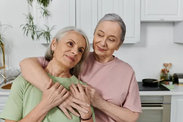 Pareja lesbiana anciana en la cocina moderna; una mujer mira hacia fuera mientras que la otra la abraza por detrás. - foto de stock