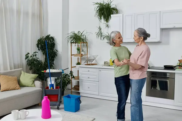 A lesbian couple in their home, one cleaning and the other standing nearby. They are both dressed casually and have grey hair. — Stock Photo