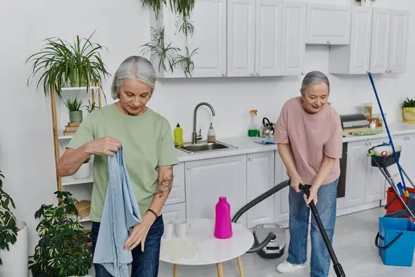 Duas mulheres maduras, um casal de lésbicas, a limpar o seu apartamento moderno. Uma mulher está aspirando enquanto a outra dobra a roupa. — Fotografia de Stock