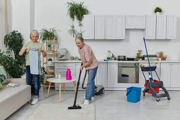 A mature lesbian couple is cleaning their modern apartment together. One woman is folding laundry while the other vacuums the floor. — Stock Photo