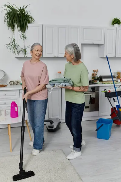 Two mature women, a lesbian couple, clean their modern apartment together. One woman vacuums while the other holds folded laundry. — Stock Photo
