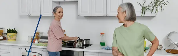 Duas mulheres maduras, um casal de lésbicas, limpam juntos o apartamento moderno. Uma mulher lava pratos enquanto a outra assiste. — Fotografia de Stock