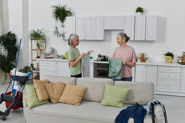 Um casal lésbico maduro limpa seu apartamento moderno juntos, uma mulher segurando uma toalha verde. — Fotografia de Stock