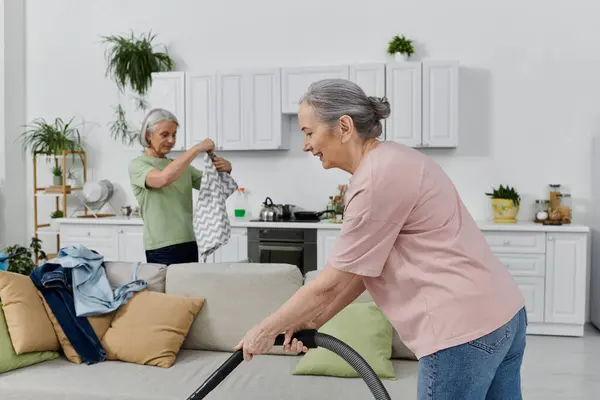Лесбійська пара чистить свою сучасну квартиру разом. Одна жінка вакуумує диван, а інша складає білизну. — стокове фото