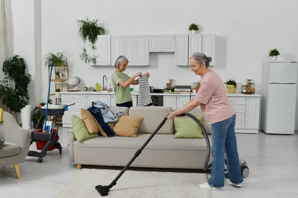 Um casal de lésbicas limpa o seu apartamento moderno juntos. Uma mulher está aspirando o chão enquanto a outra dobra a roupa. — Fotografia de Stock