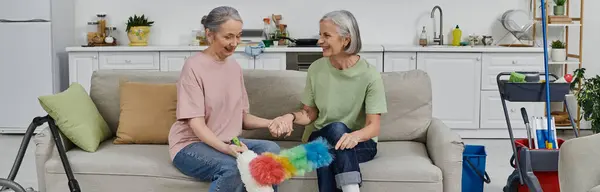 A mature lesbian couple cleans their modern apartment together, with one woman holding a feather duster. — Stock Photo