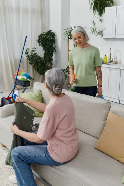 Zwei ältere lesbische Frauen putzen ihre Wohnung, eine auf dem Sofa, die andere steht und hält eine Sprühflasche in der Hand. — Stockfoto