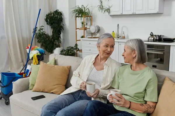 A mature lesbian couple relaxes on their couch, enjoying a coffee break after cleaning their modern apartment. — Stock Photo