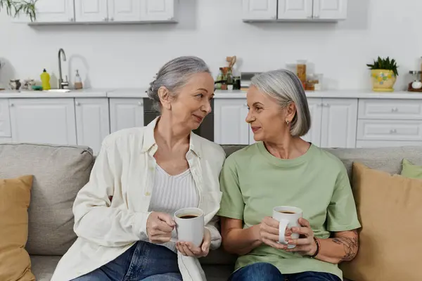 Two mature lesbian women relax on a couch in their modern apartment, enjoying coffee after a cleaning session. — Stock Photo