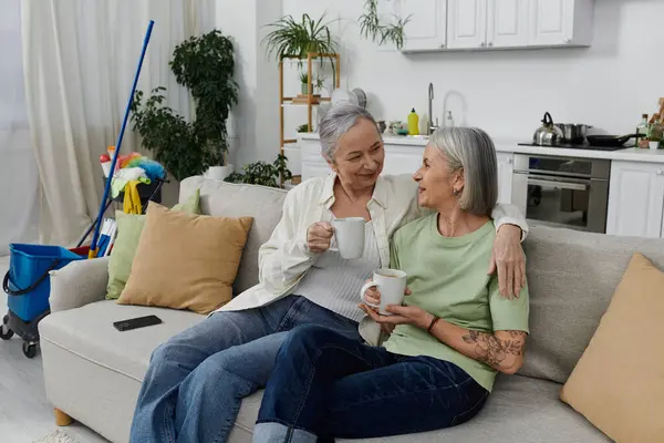 Un couple de lesbiennes mûres se détend sur le canapé dans leur appartement moderne, dégustant du café après une journée de nettoyage. — Photo de stock