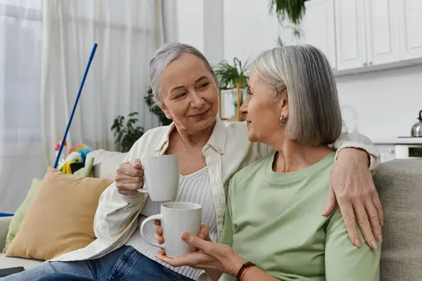 Duas mulheres lésbicas maduras relaxam em um sofá em seu apartamento moderno após a limpeza, desfrutando de uma bebida quente e companhia uns dos outros.. — Fotografia de Stock