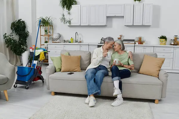 A mature lesbian couple relaxes on the couch after cleaning their modern apartment together. — Stock Photo