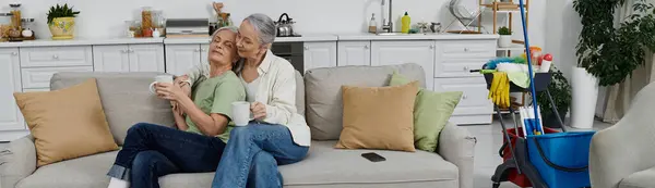 Two mature lesbian women relaxing on a couch in their modern apartment after cleaning together. — Stock Photo