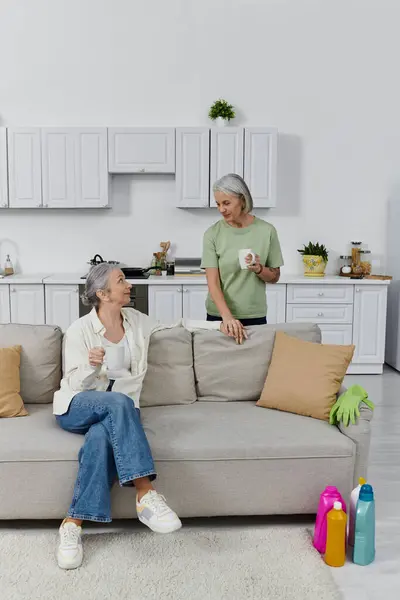 A mature lesbian couple relaxes on a couch in their modern apartment after cleaning. — Stock Photo