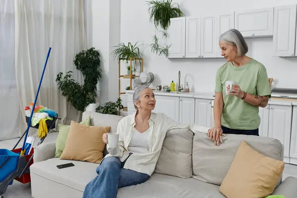 Two mature lesbian women relax on a couch in their modern apartment after finishing cleaning. They enjoy their coffee and conversation. — Stock Photo