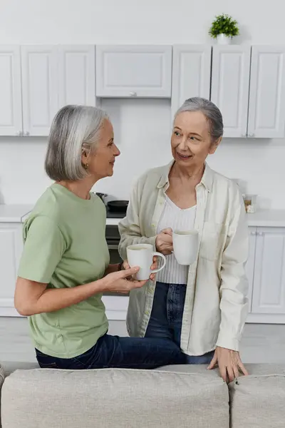 A mature lesbian couple relaxes with coffee after cleaning their modern apartment. — Stock Photo