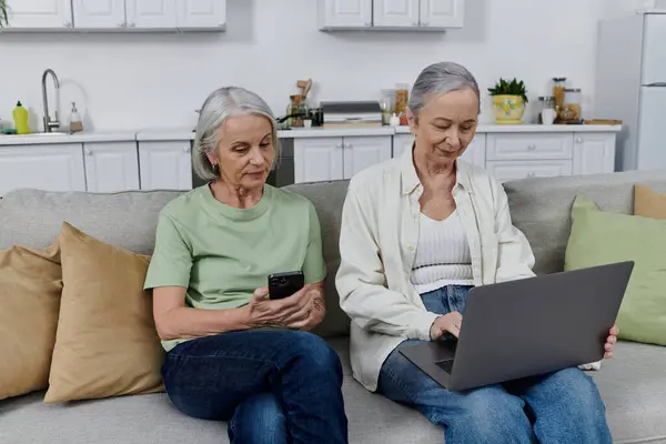 Dos mujeres felices, una pareja lesbiana, se detienen de ordenar su apartamento moderno: una mira a su teléfono, la otra trabaja en una computadora portátil. - foto de stock