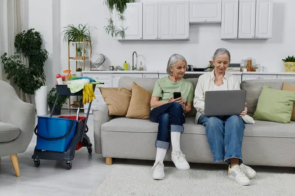 Two mature lesbians relax on a couch in their modern apartment. One on her phone, the other on her laptop. — Stock Photo