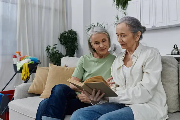 Zwei reife lesbische Frauen entspannen sich auf einer Couch in ihrer modernen Wohnung und lesen ein Buch, nachdem sie ihre Putzarbeiten erledigt haben. — Stockfoto