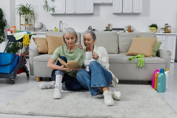 A mature lesbian couple relaxes on the floor of their modern apartment, enjoying a book and a cup of coffee after cleaning. — Stock Photo