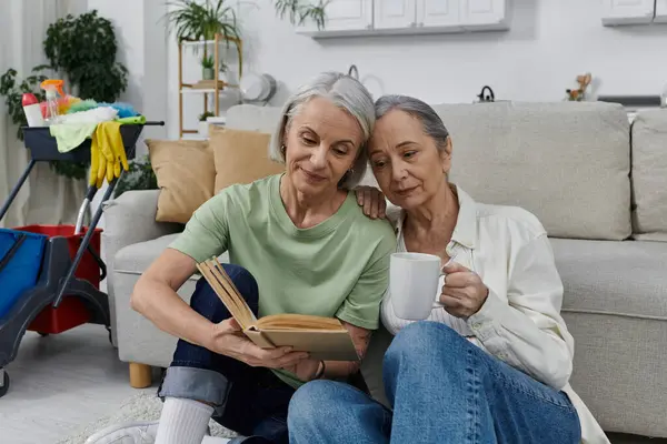 A mature lesbian couple relaxes on the floor of their modern apartment after cleaning, sharing a book and a warm drink. — Stock Photo