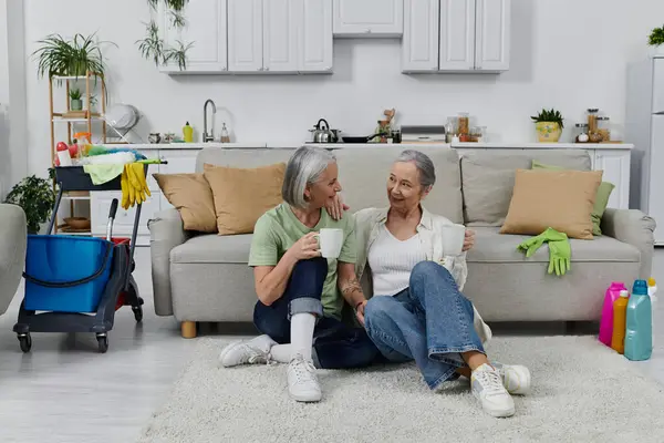 Two mature lesbian women rest on a rug in their modern apartment after finishing cleaning, enjoying a cup of tea. — Stock Photo