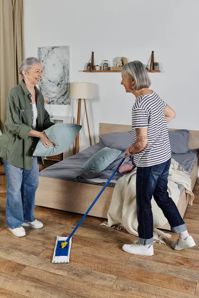 Duas mulheres lésbicas maduras limpam seu apartamento moderno juntas, uma fazendo a cama enquanto a outra esfrega o chão. — Fotografia de Stock